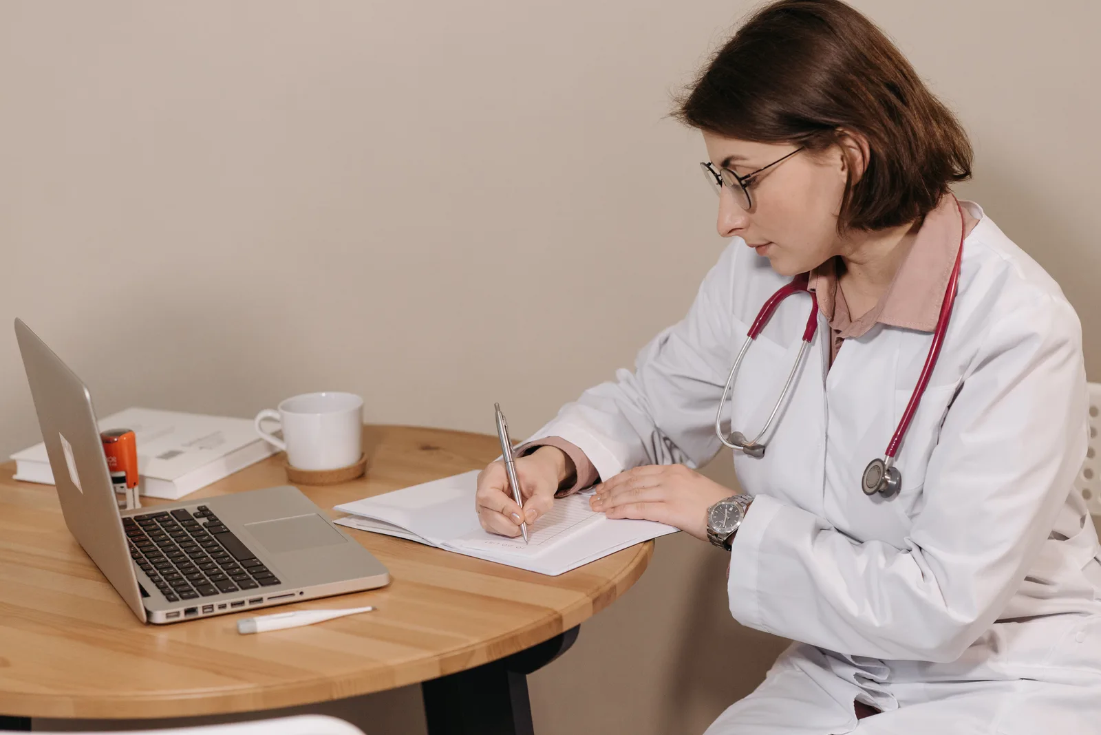 A clinician writing notes in a notebook beside an open laptop.